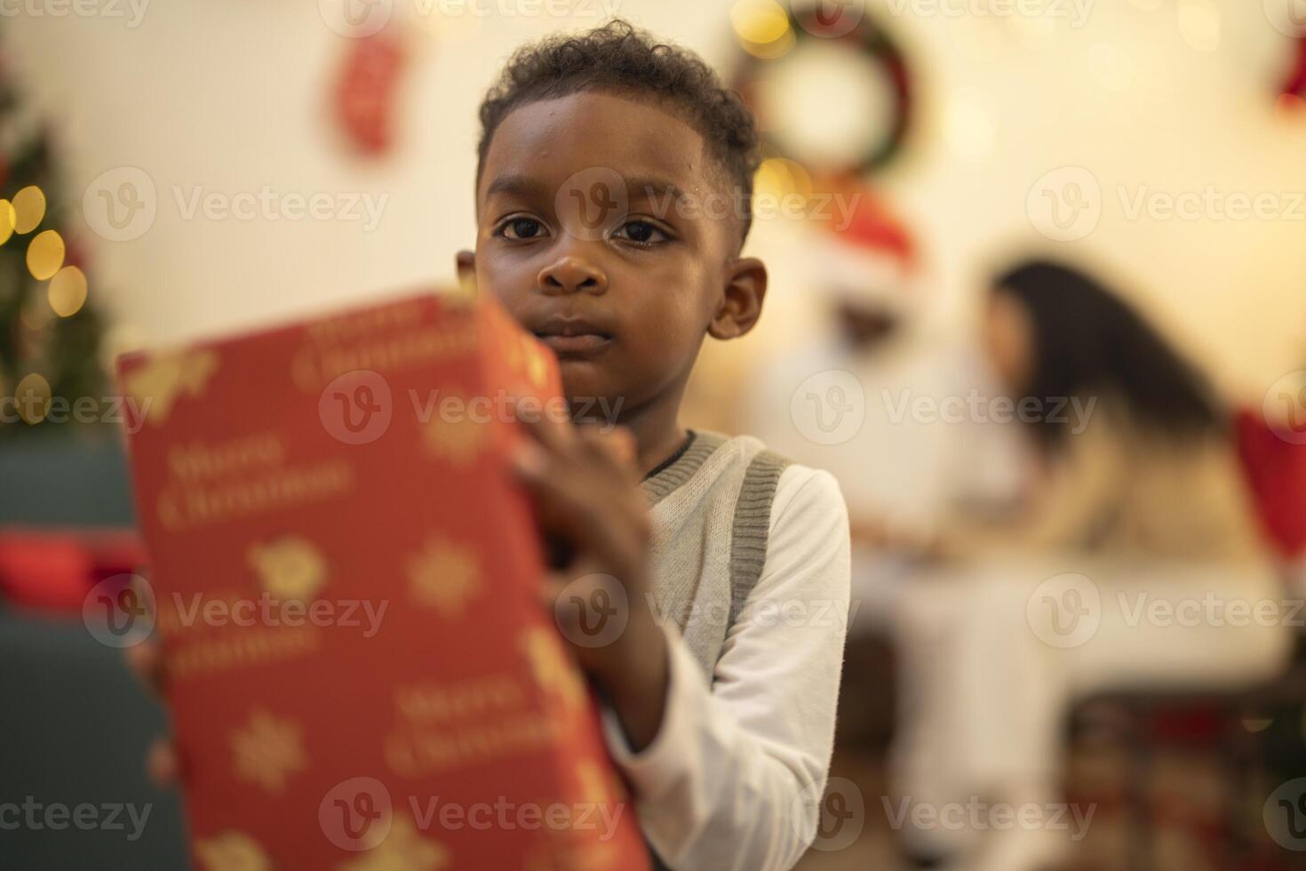 A young child excitedly holding a Christmas gift box, surrounded by festive decorations and twinkling lights in the background. The joyful expression on the child's face reflects the happiness. photo