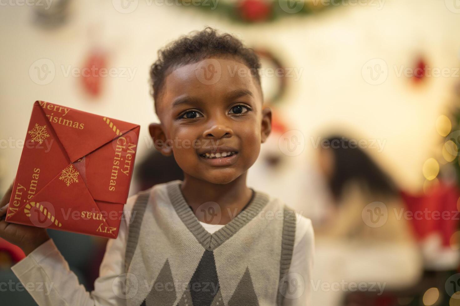 A young child excitedly holding a Christmas gift box, surrounded by festive decorations and twinkling lights in the background. The joyful expression on the child's face reflects the happiness. photo