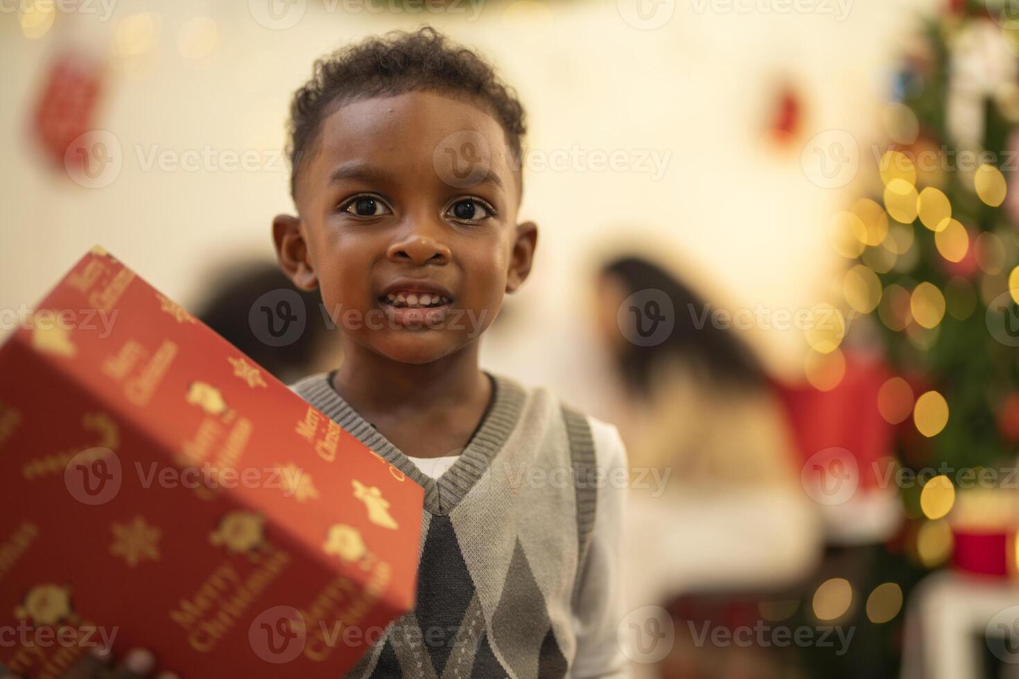 A young child excitedly holding a Christmas gift box, surrounded by festive decorations and twinkling lights in the background. The joyful expression on the child's face reflects the happiness. photo