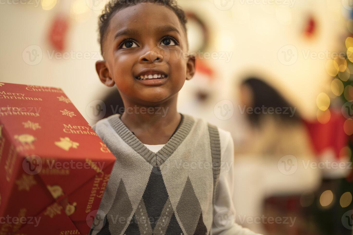 A young child excitedly holding a Christmas gift box, surrounded by festive decorations and twinkling lights in the background. The joyful expression on the child's face reflects the happiness. photo