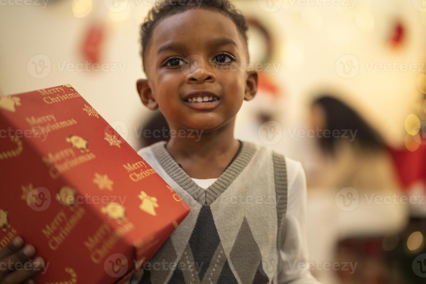 A young child excitedly holding a Christmas gift box, surrounded by festive decorations and twinkling lights in the background. The joyful expression on the child's face reflects the happiness. photo