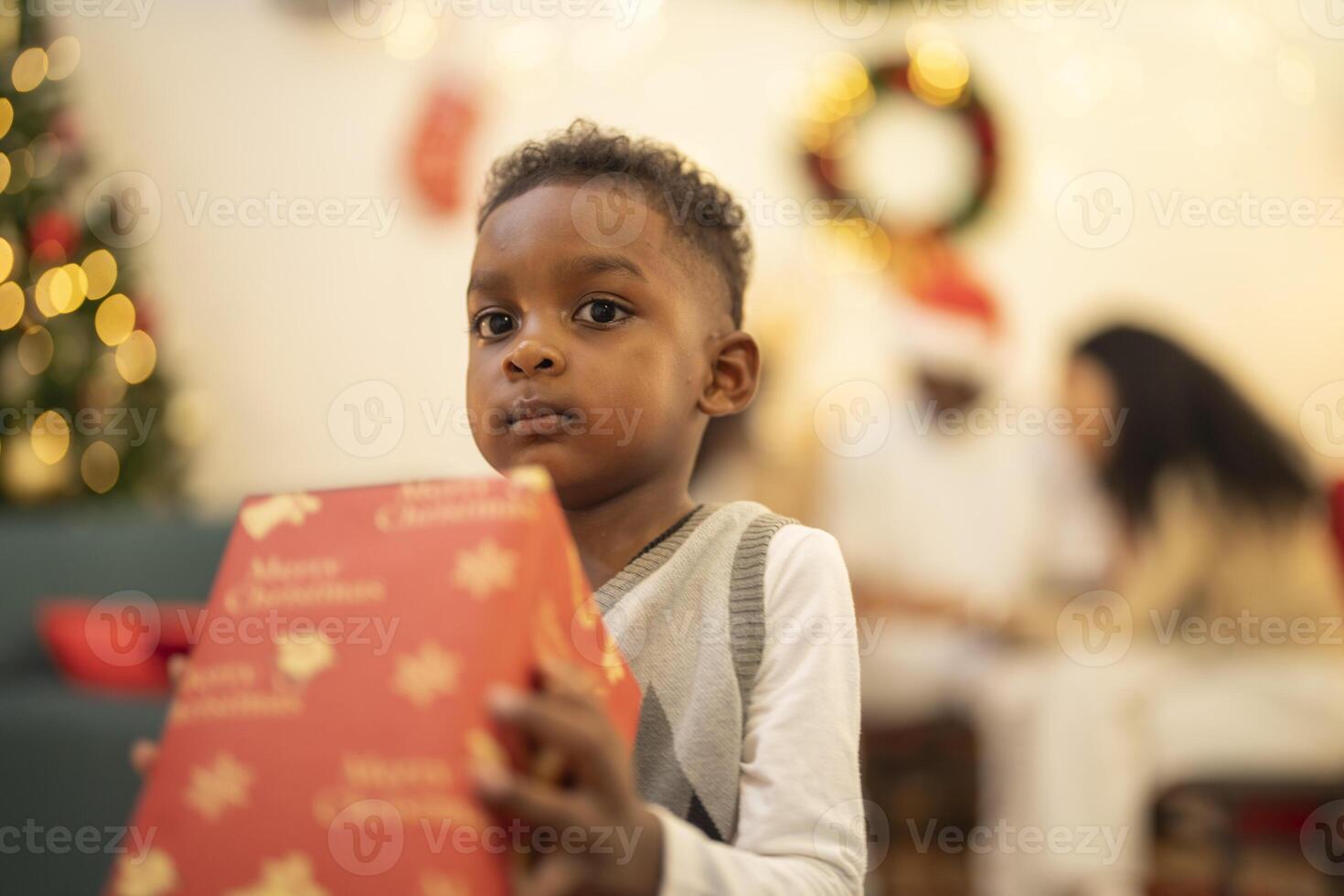 A young child excitedly holding a Christmas gift box, surrounded by festive decorations and twinkling lights in the background. The joyful expression on the child's face reflects the happiness. photo