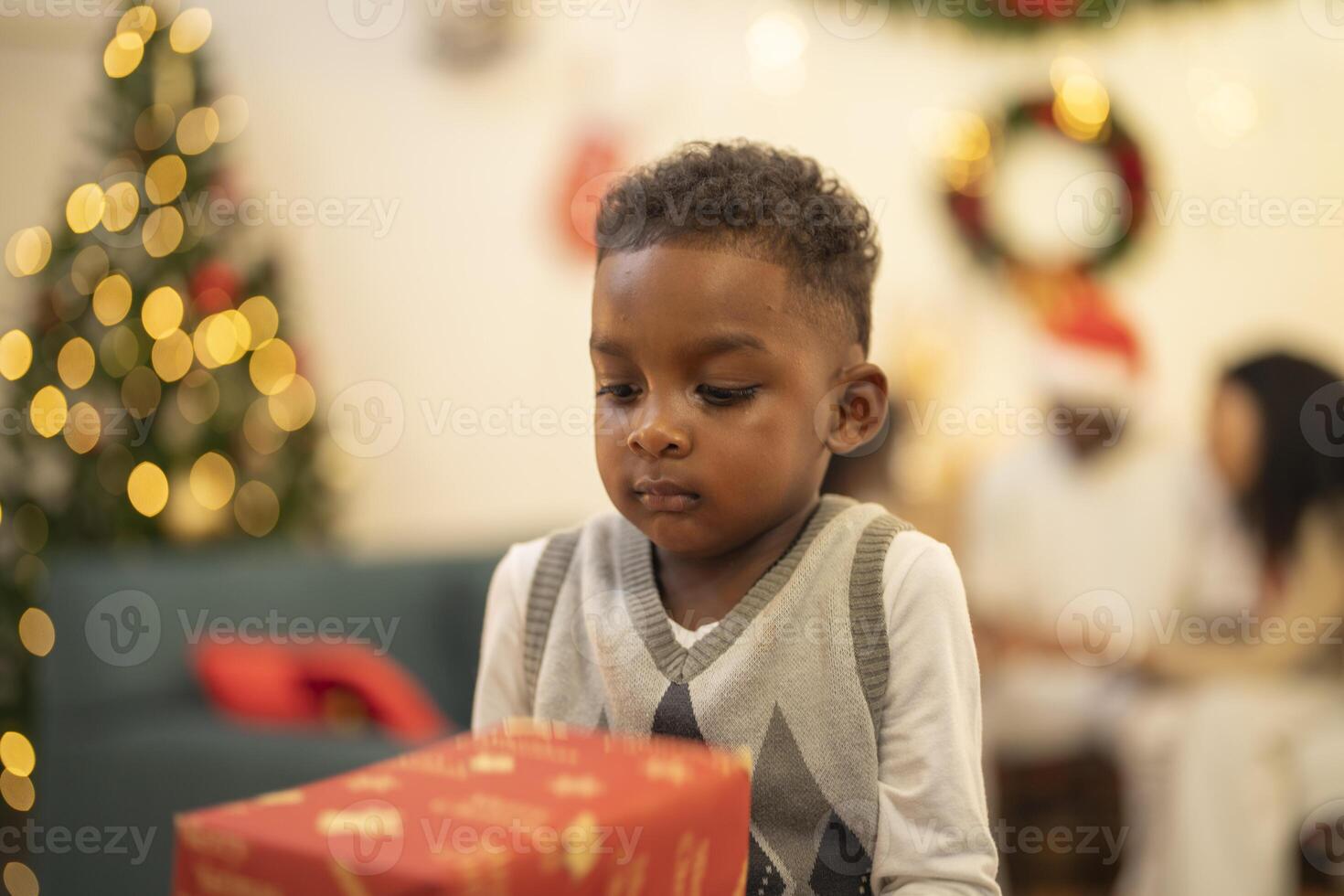 A young child excitedly holding a Christmas gift box, surrounded by festive decorations and twinkling lights in the background. The joyful expression on the child's face reflects the happiness. photo