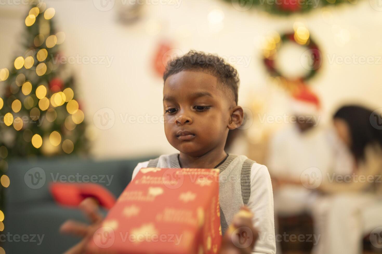 A young child excitedly holding a Christmas gift box, surrounded by festive decorations and twinkling lights in the background. The joyful expression on the child's face reflects the happiness. photo