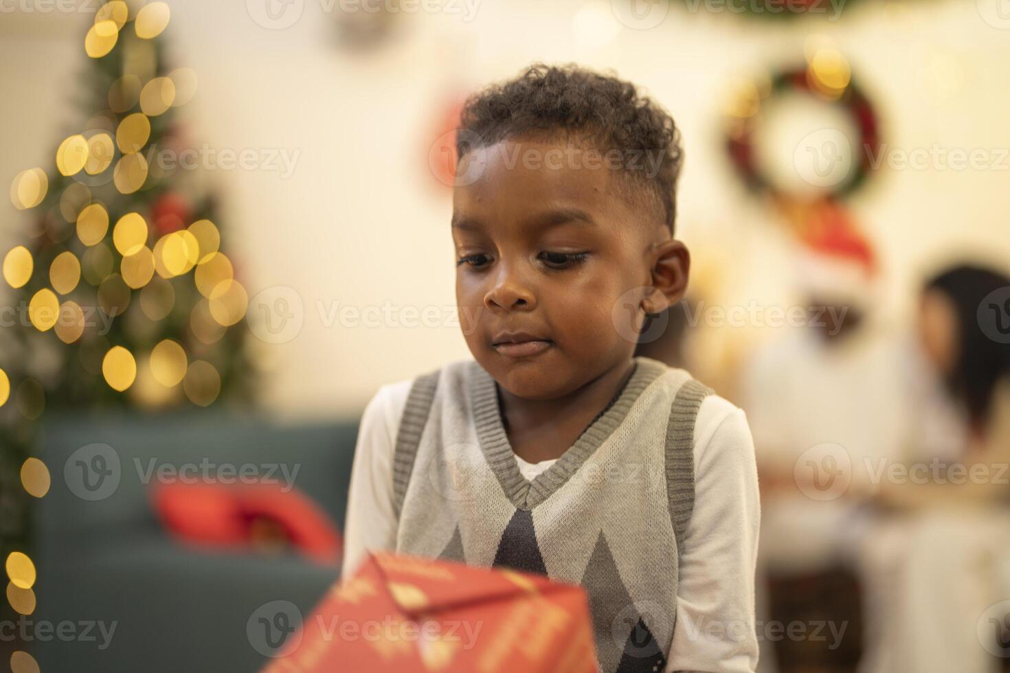 A young child excitedly holding a Christmas gift box, surrounded by festive decorations and twinkling lights in the background. The joyful expression on the child's face reflects the happiness. photo