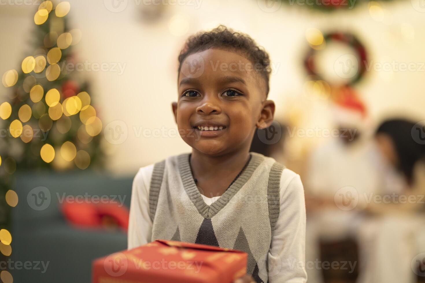 A young child excitedly holding a Christmas gift box, surrounded by festive decorations and twinkling lights in the background. The joyful expression on the child's face reflects the happiness. photo