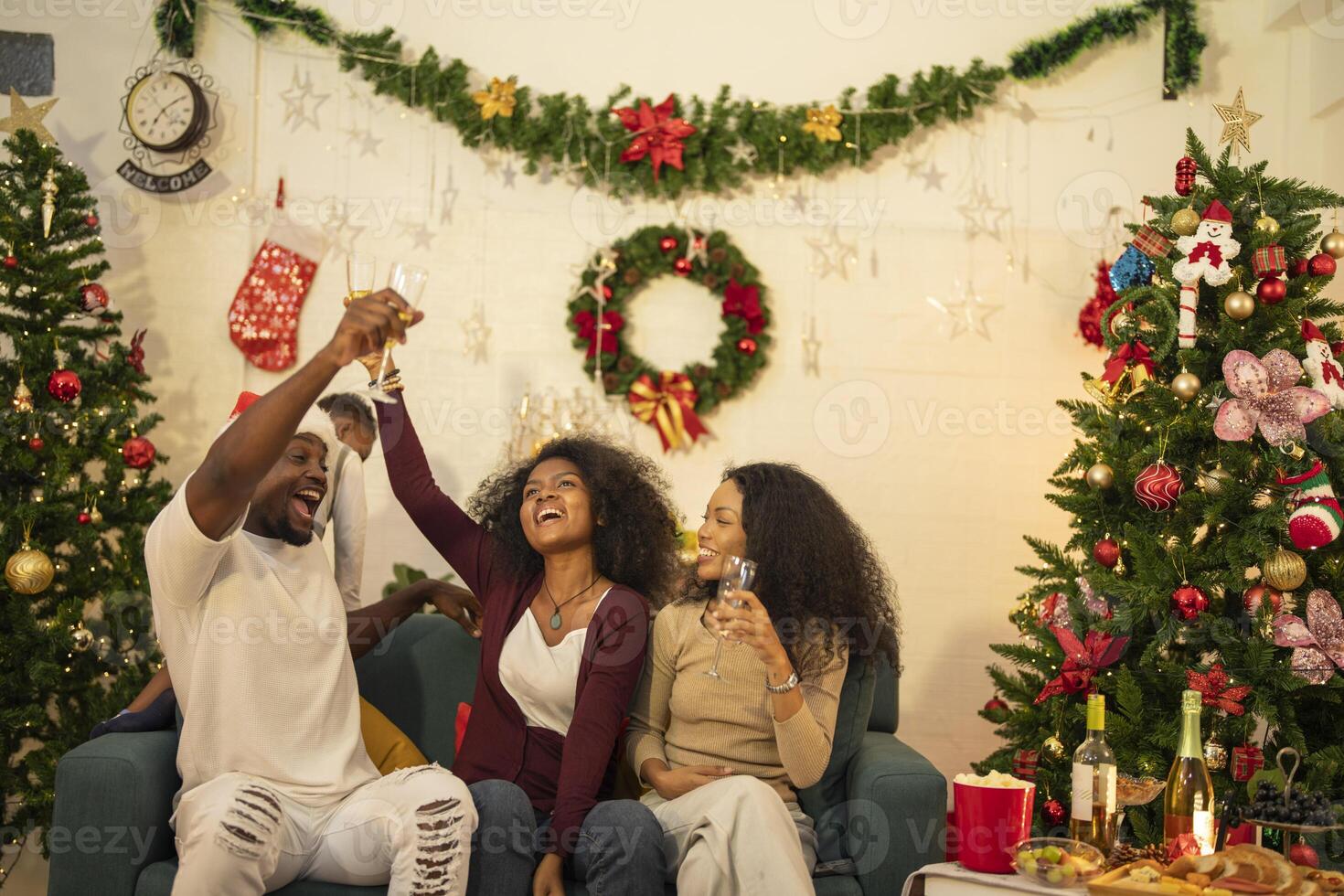 A lively Christmas celebration with three african friends enjoying drinks and holiday cheer on a sofa. The festive atmosphere includes Christmas trees, wreaths, lights, and decorations. photo