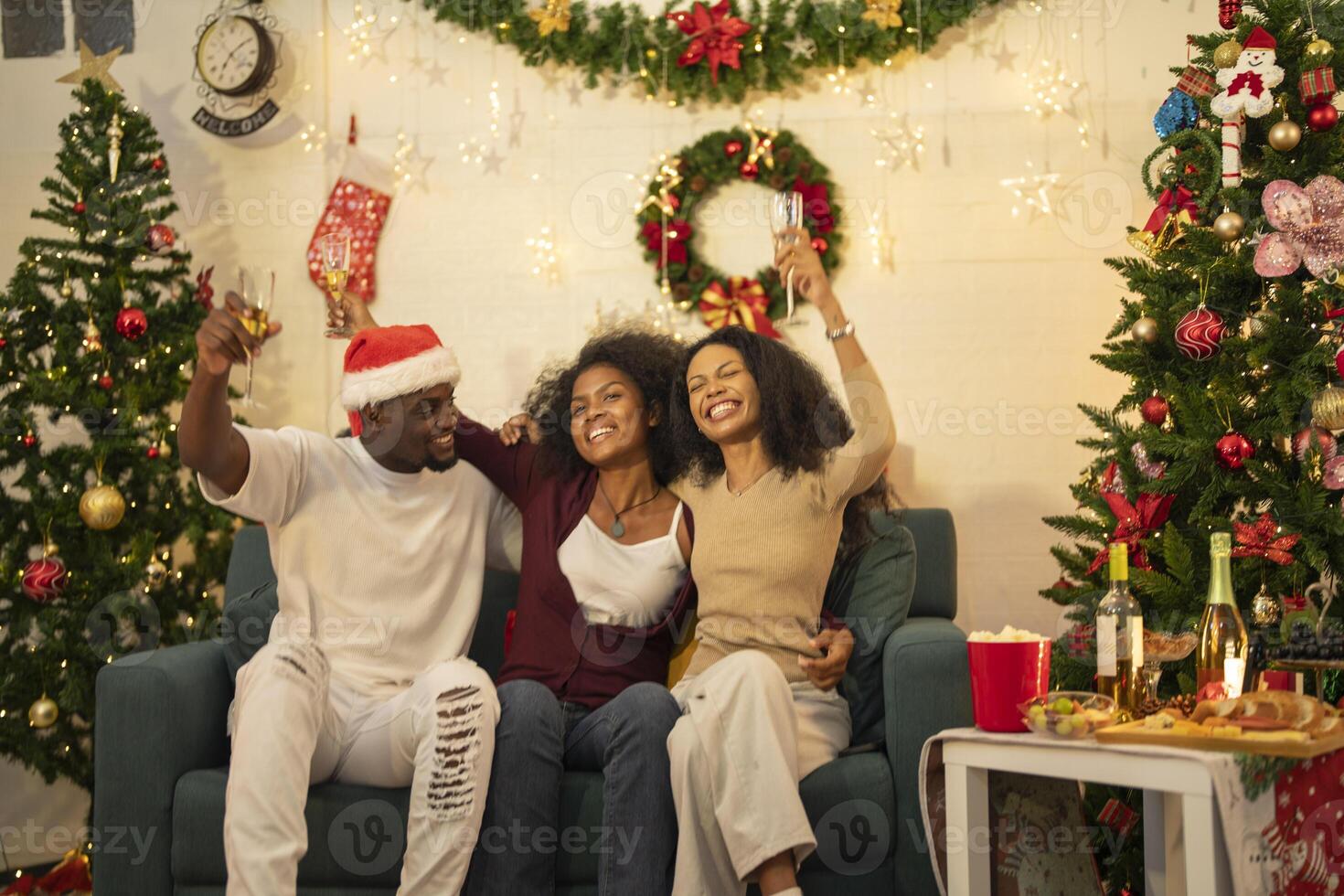 A lively Christmas celebration with three african friends enjoying drinks and holiday cheer on a sofa. The festive atmosphere includes Christmas trees, wreaths, lights, and decorations. photo