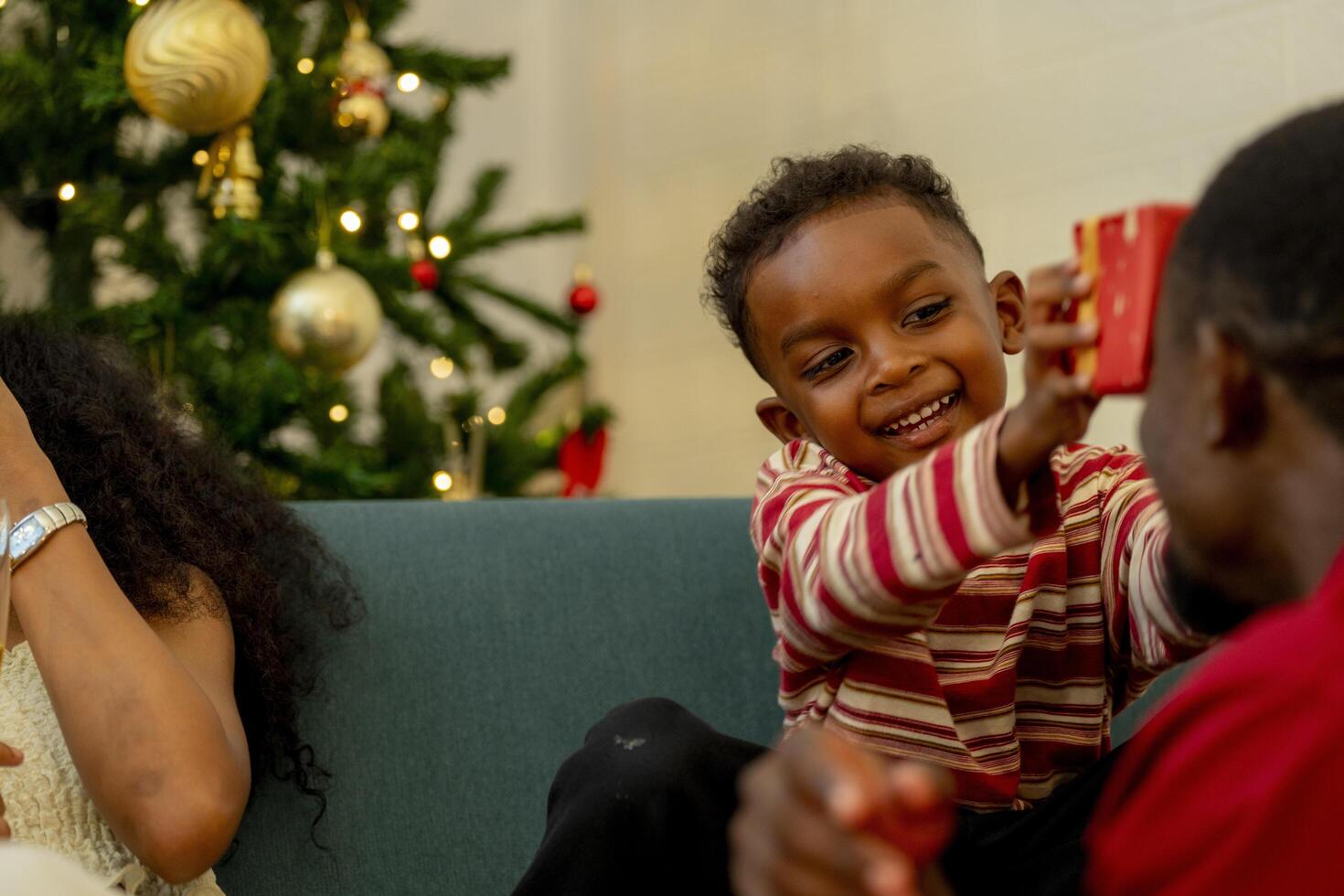 A cheerful family gathers around the Christmas tree, sharing a moment of joy as a man presents a red gift box to a child. while family members relax on the sofa, enjoying the warmth of the season. photo