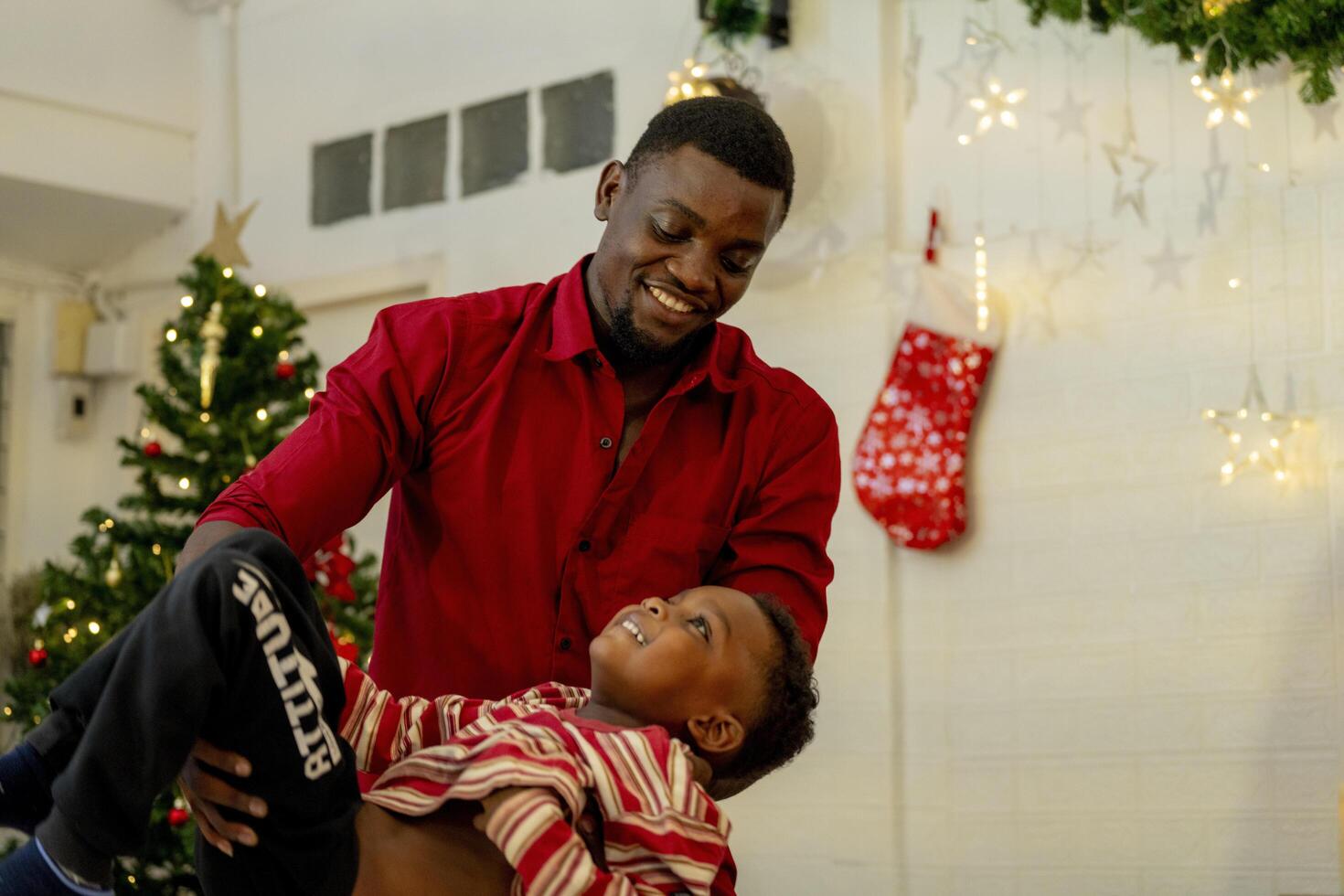 A joyful father plays with his son near a beautifully decorated Christmas tree. The room is filled with holiday cheer, featuring twinkling Christmas lights, stockings, and festive decorations. photo