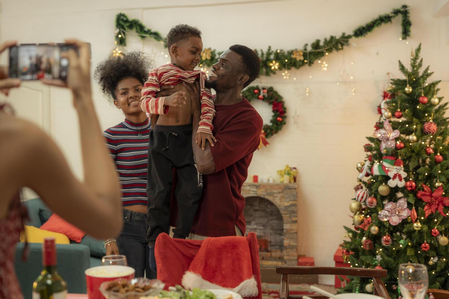 A joyful family gathers around the dinner table celebrating Christmas. The father, dressed in a Santa hat, gives a gift to the child, while everyone smiles with warmth and festive decorations. photo