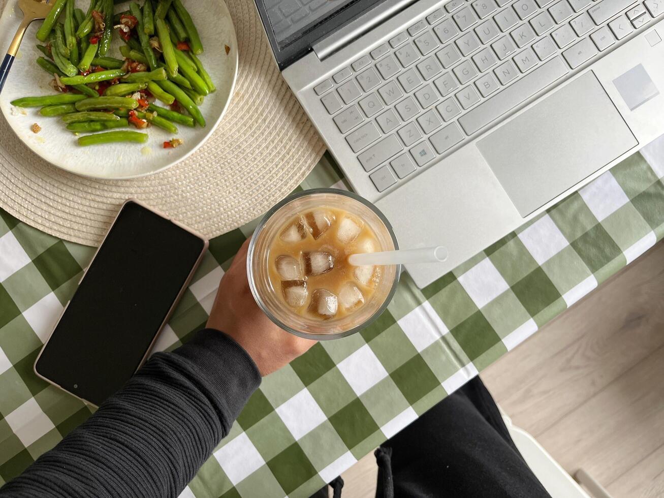 A hand holding a glass of iced coffee with Laptop and green beans on the table photo