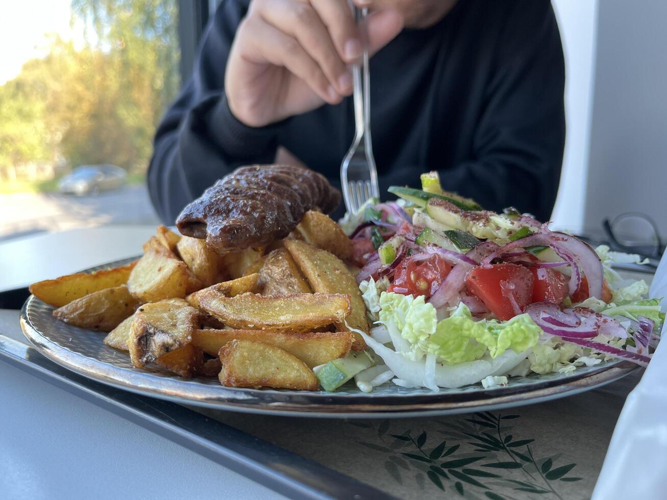 Unrecognized man holding plastic fork for enjoying Kebab with French fries and salad on a plate on white table in a restaurant photo
