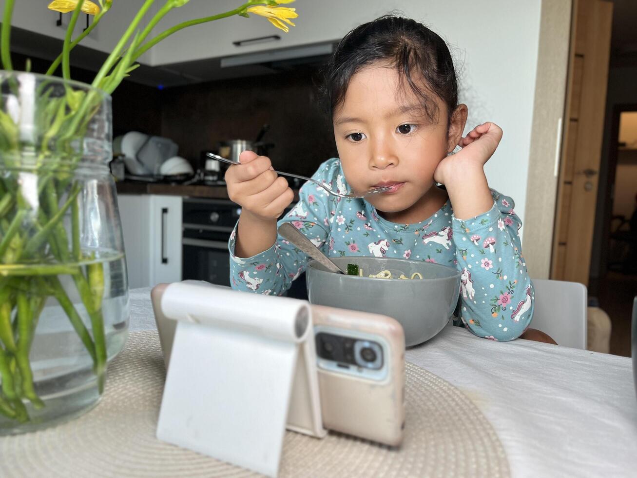 Asian little girl watching mobile phone while eating in the modern kitchen. Screen time health risks concept photo