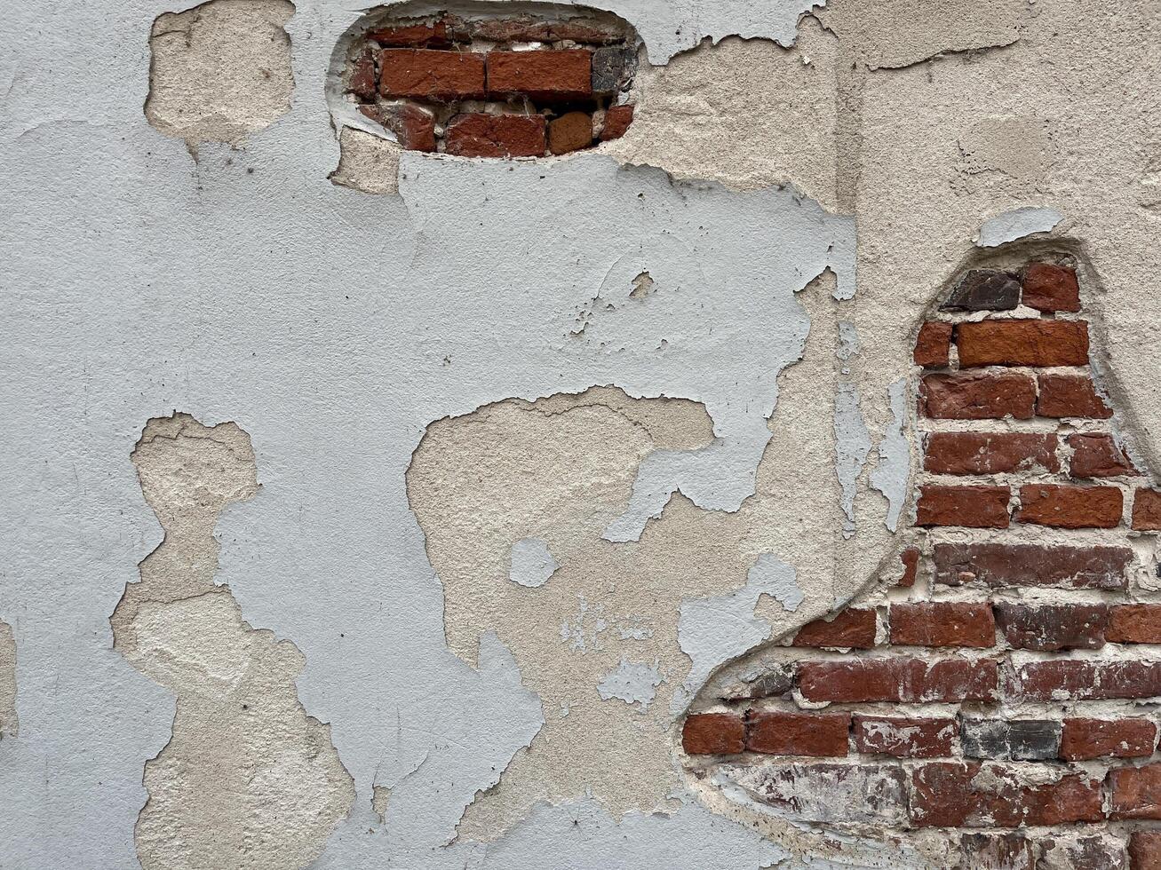 A weathered wall with peeling white plaster, revealing patches of red brick underneath. The plaster is cracked and chipped in several places, creating a textured and aged appearance. photo