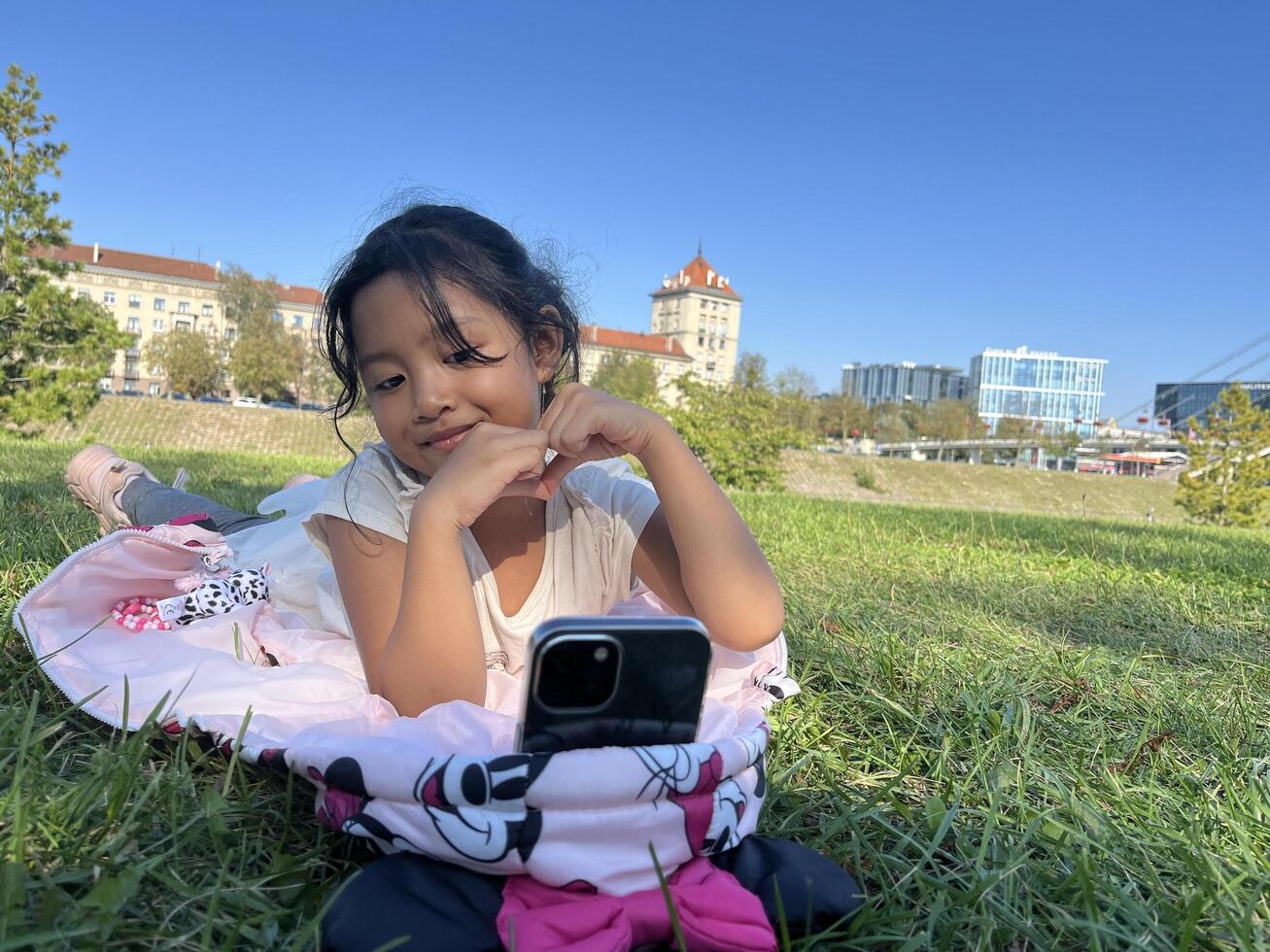Smiling Asian little girl on the grass using phone in outdoor making call with love sign hand gesture photo