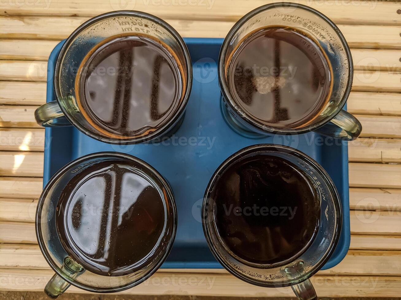 Top-down view of four cups filled with black coffee placed on a blue tray. The reflective surface of the coffee contrasts with the transparent glass mugs. . photo