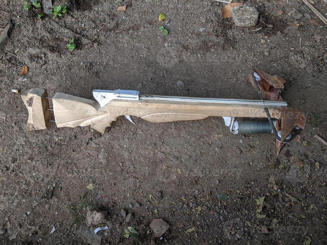 A handcrafted wooden crossbow with a visible string and trigger mechanism, lying on a dirt ground. The crossbow has a rustic, homemade appearance. photo
