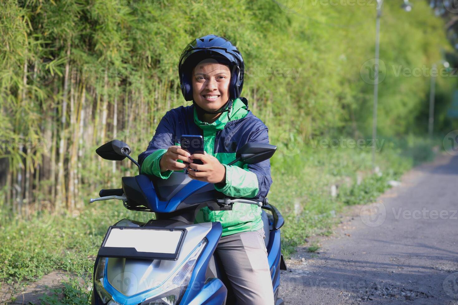 Online taxi rider wearing green jacket and helmet, riding motorcycle, using a mobile phone while on the way to pick up customer. Transportation application concept. photo