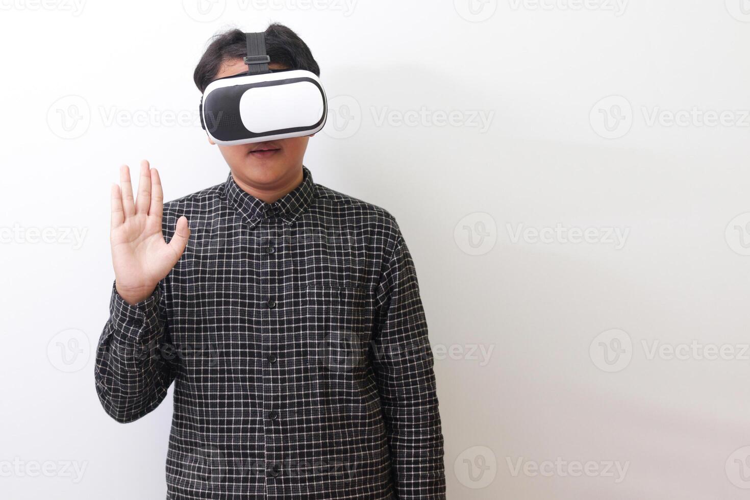 Portrait of Asian man in black plaid shirt using Virtual Reality VR glasses and greet his friend inside the game with opened palm. Isolated image on white background photo