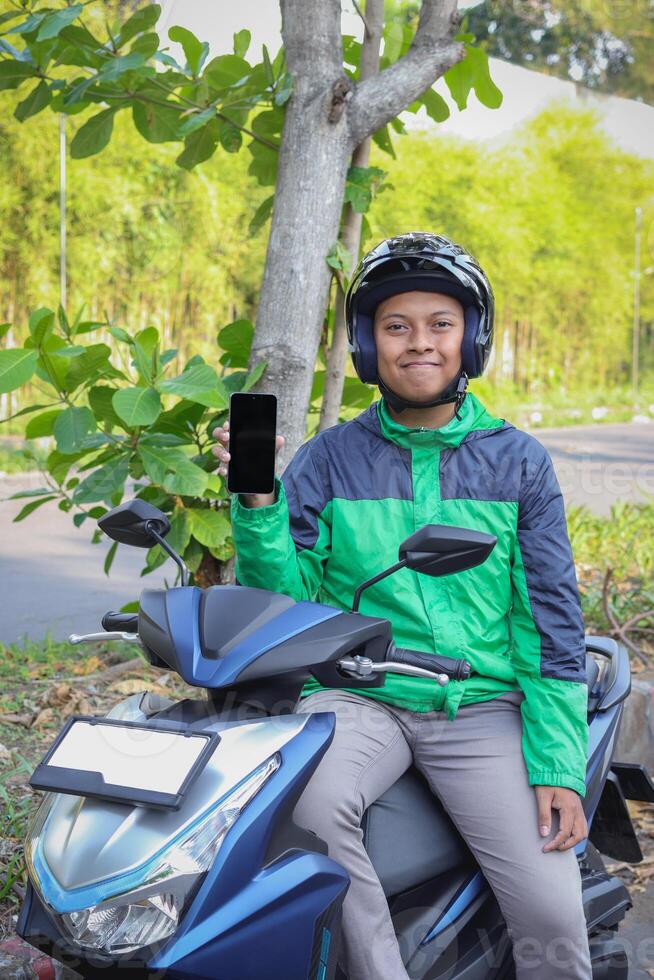 Online taxi rider wearing green jacket and helmet, riding motorcycle, showing blank screen mobile phone while on the way to pick up customer. Transportation application concept. Smartphone mockup photo