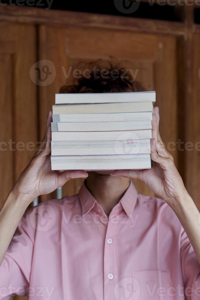 Mysterious Asian man covering his face with a stack of books. Education and knowledge concept. Portrait of young male book lover photo