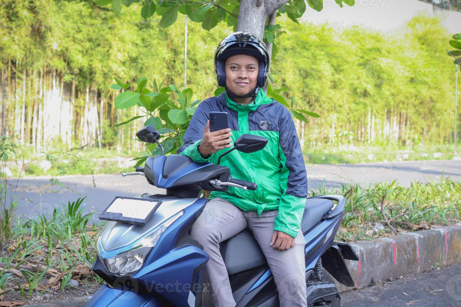 Online taxi rider wearing green jacket and helmet, riding motorcycle, using a mobile phone while on the way to pick up customer. Transportation application concept. photo