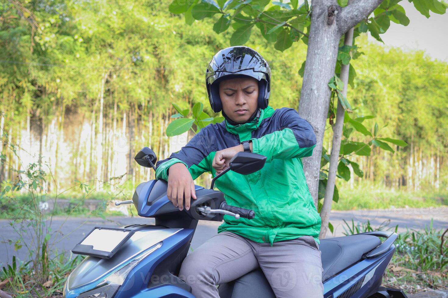 Online taxi rider wearing green jacket and helmet, riding motorcycle, checking time on his watch while on the way to pick up customer. Transportation application concept. photo