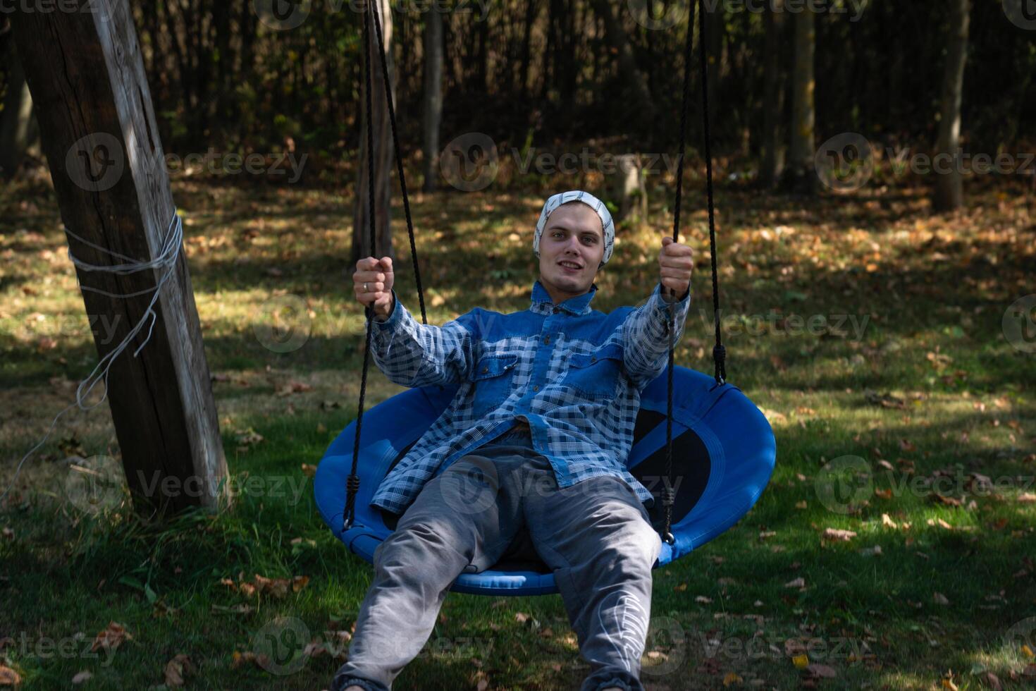 A cheerful young guy riding a swing in the village. A guy in a shirt and a scarf photo