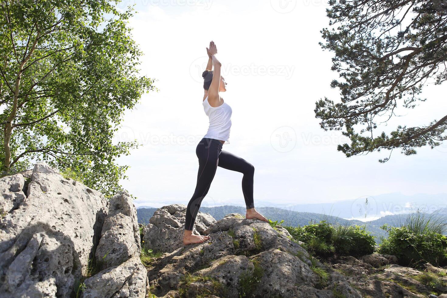 Attractive young woman doing a yoga pose for balance and stretching staying on top of high rock in the mountains photo