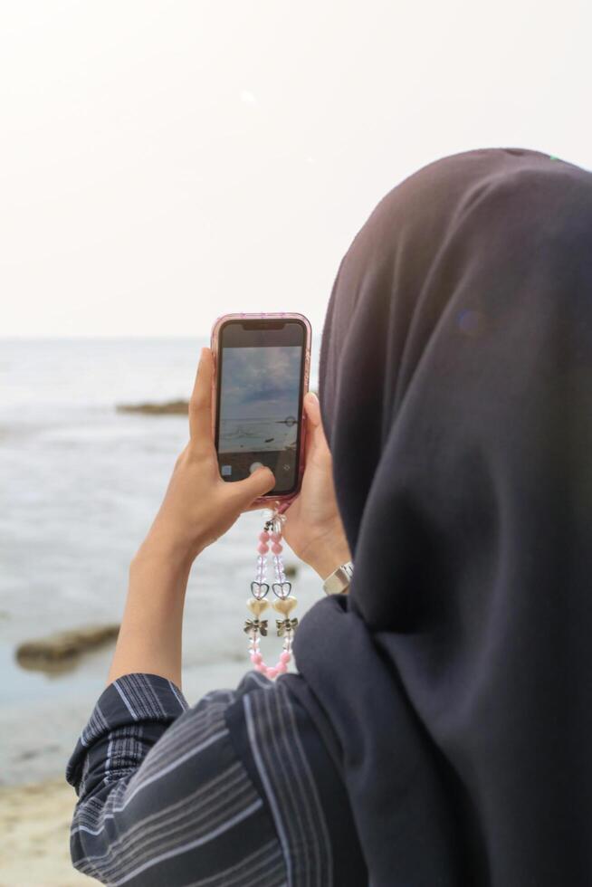 A woman wearing a hijab taking a photo of the ocean