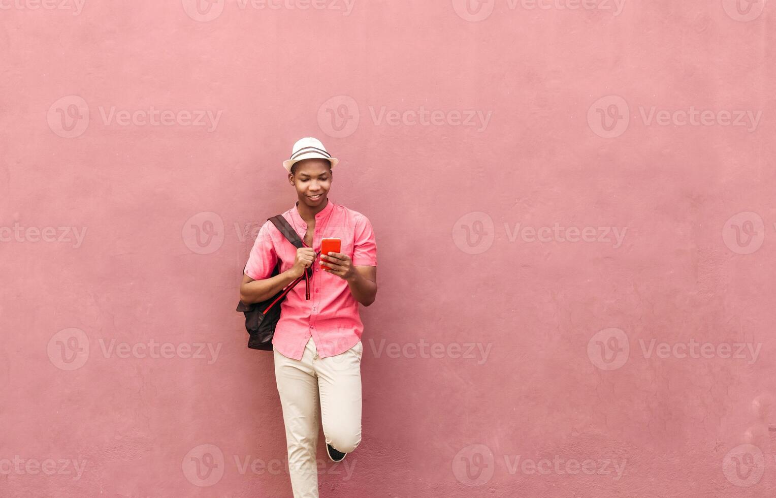Latin american man with backpack and hat stand over a colorful wall looking a smartphone mobile. photo