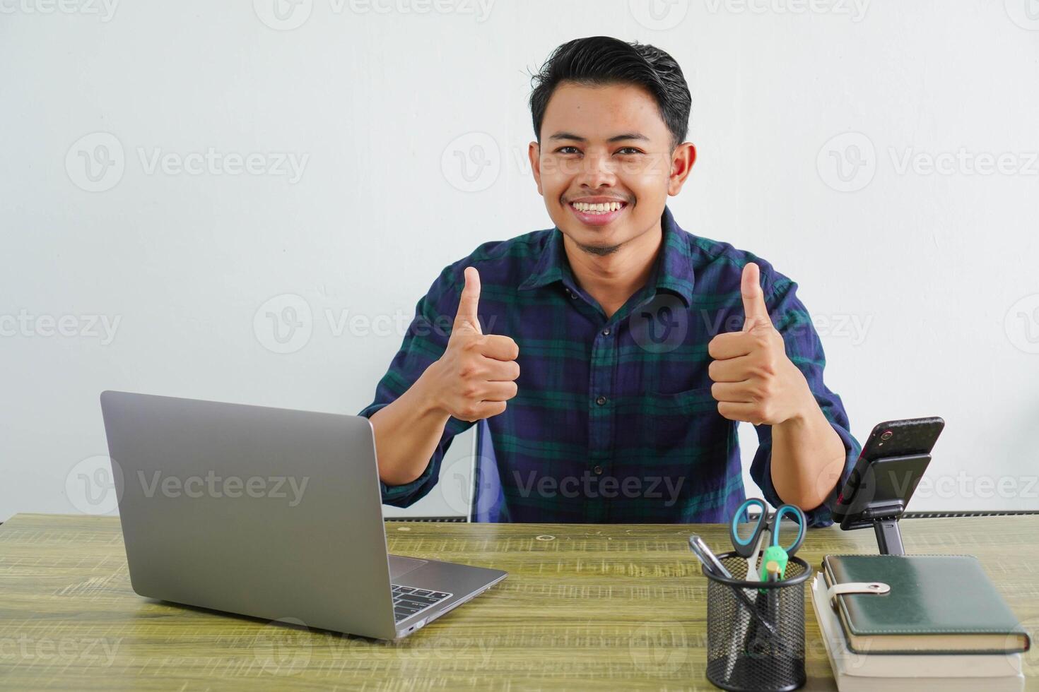 Smiling young asian man in blue shirt sit work at wooden desk with pc laptop isolated on white background. Achievement business career lifestyle concept. showing thumb up photo