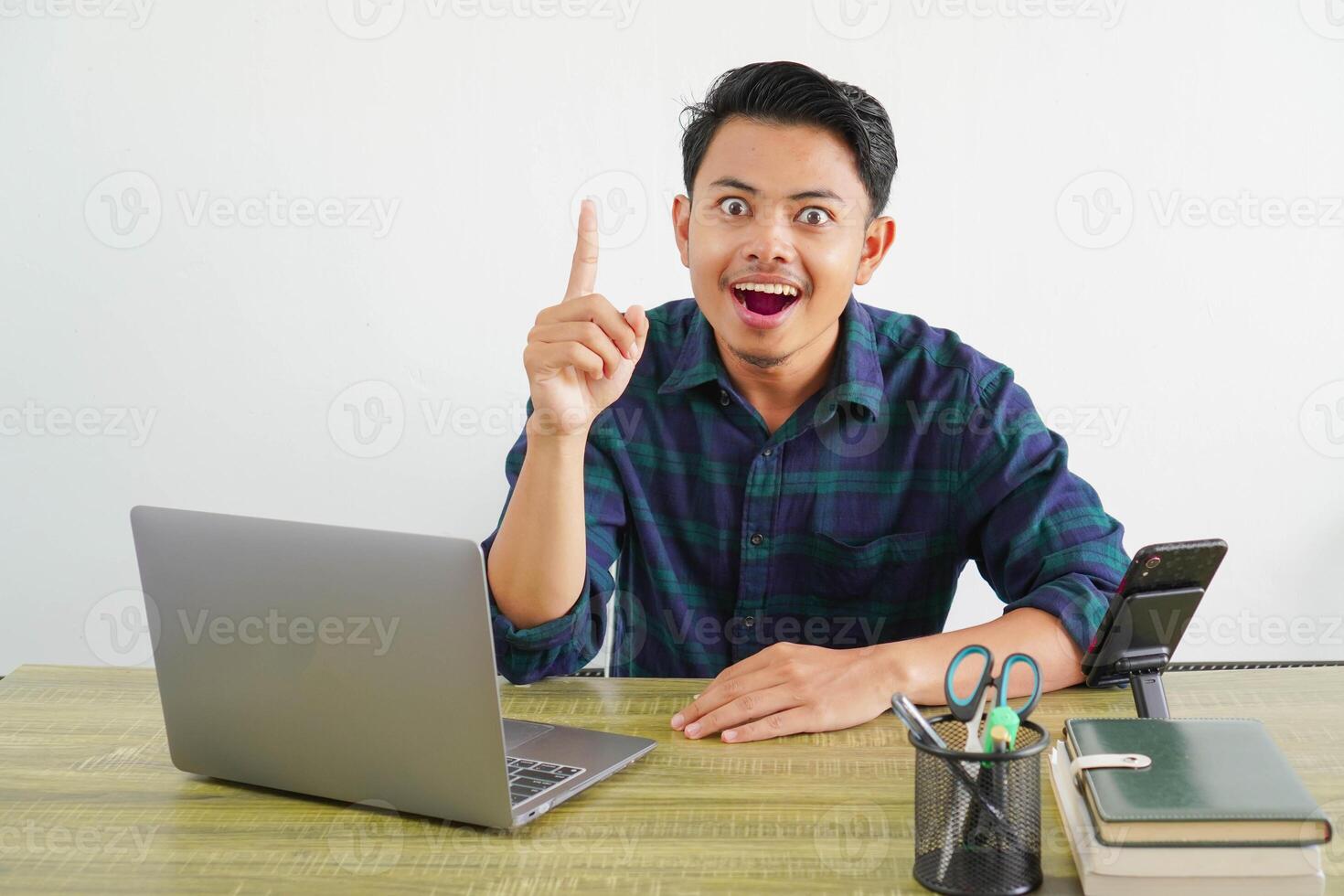 Excited young asian man sit work at wooden desk with pc laptop. Achievement business career lifestyle concept. Holding index finger up with great new idea photo