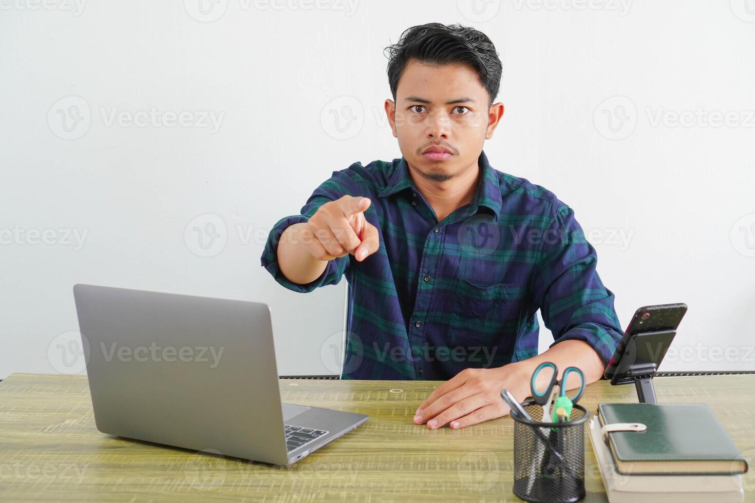 strict young asian man sit work at wooden desk with pc laptop. Achievement business career lifestyle concept. pointing index finger on camera photo