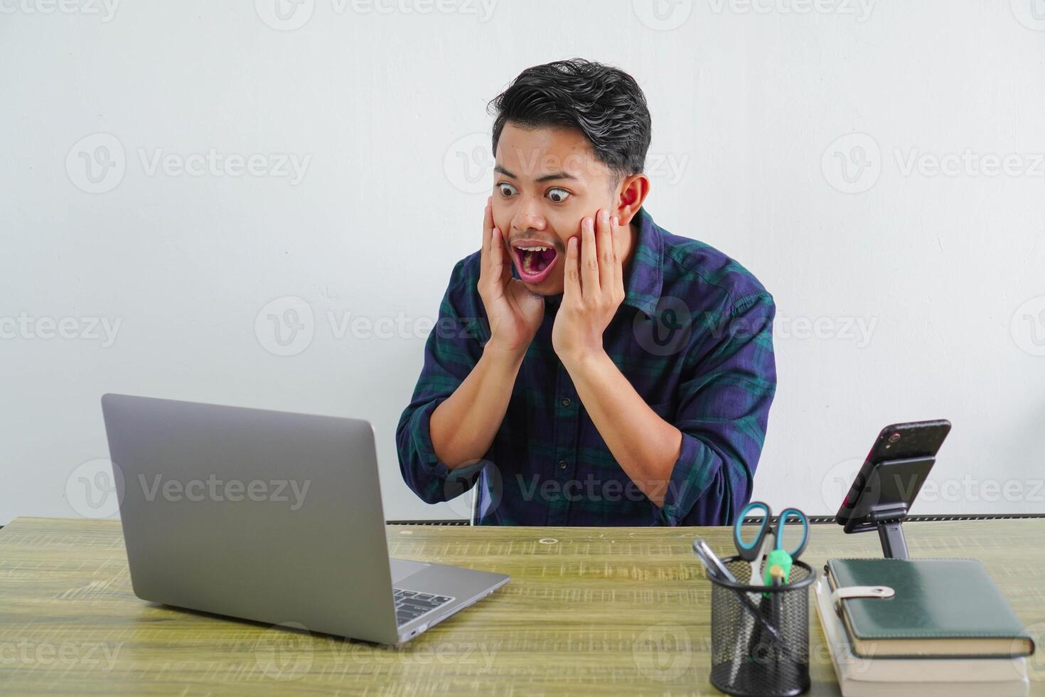 Surprised young asian man sit work at wooden desk with pc laptop. Achievement business career lifestyle concept. put hands on cheeks photo