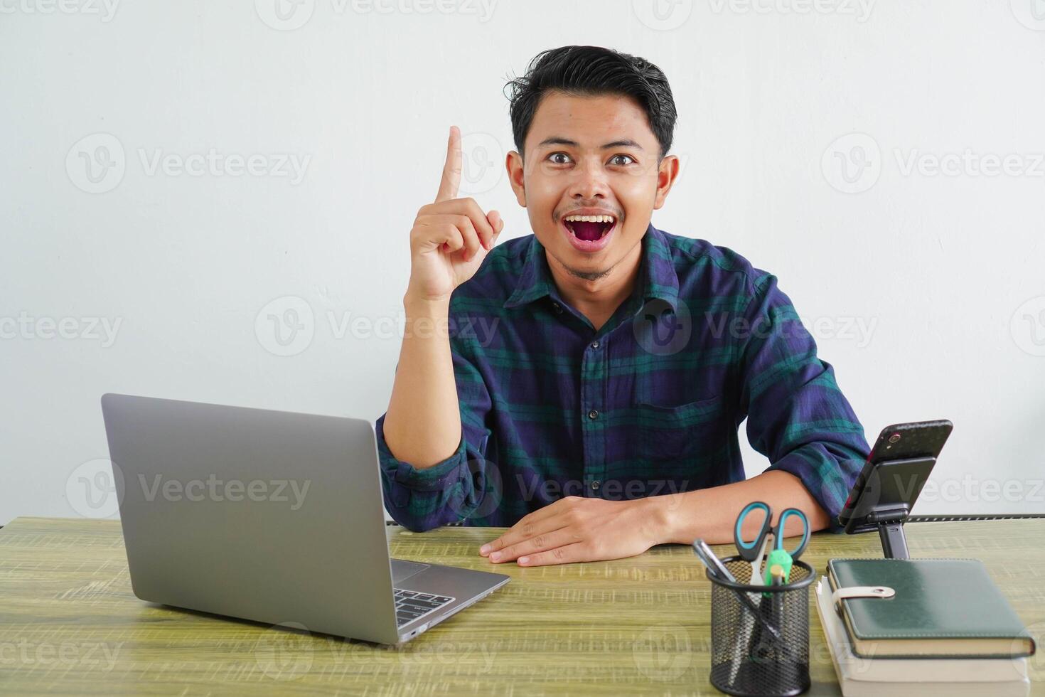 Joyful young asian man sit work at wooden desk with pc laptop. Achievement business career lifestyle concept. holding index finger up with great new idea photo