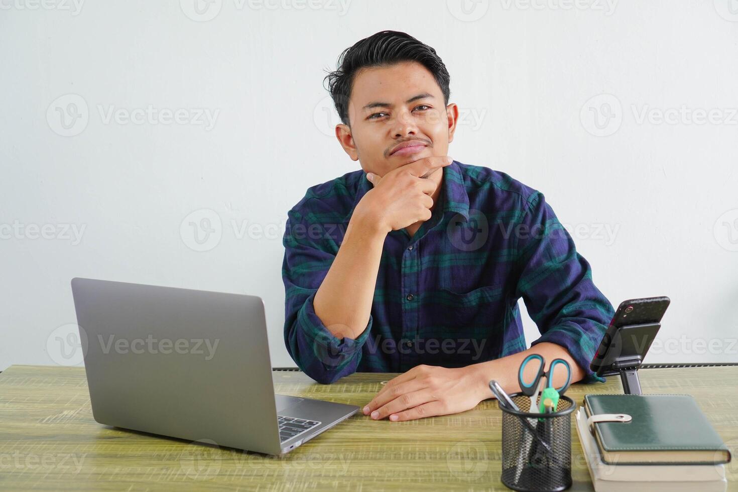 Confused young asian man in blue shirt sit work at wooden desk with pc laptop isolated on white background. Achievement business career lifestyle concept. Put hand prop up on chin photo