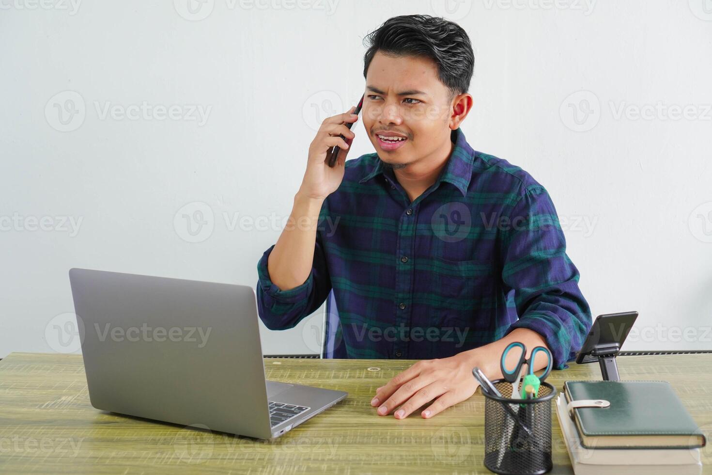 Smiling young asian man in blue shirt sit work at wooden desk with pc laptop isolated on white background. Achievement business career lifestyle concept. Talking on mobile phone photo