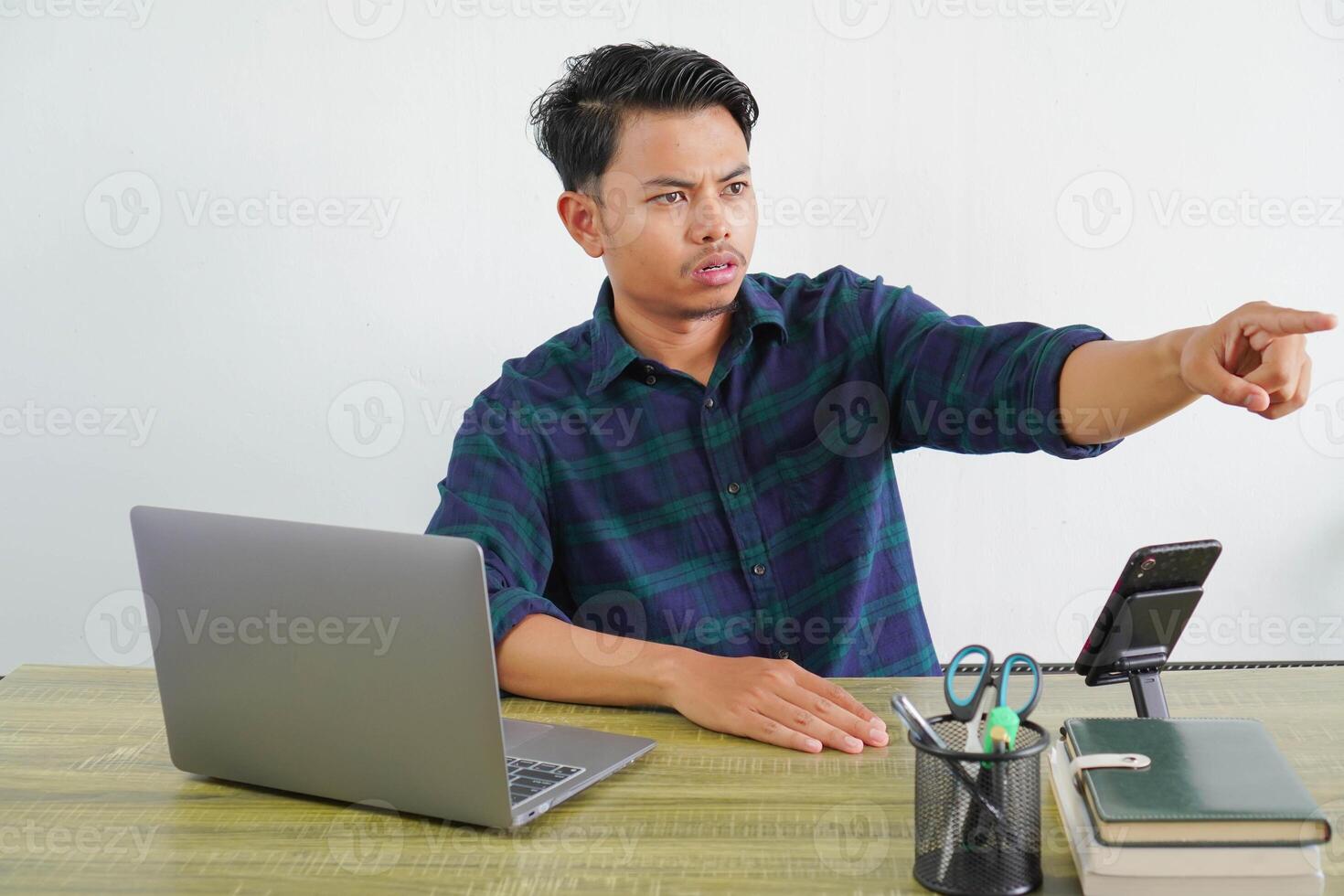 puzzled young asian man sit work at wooden desk with pc laptop. Achievement business career lifestyle concept. pointing index finger aside photo