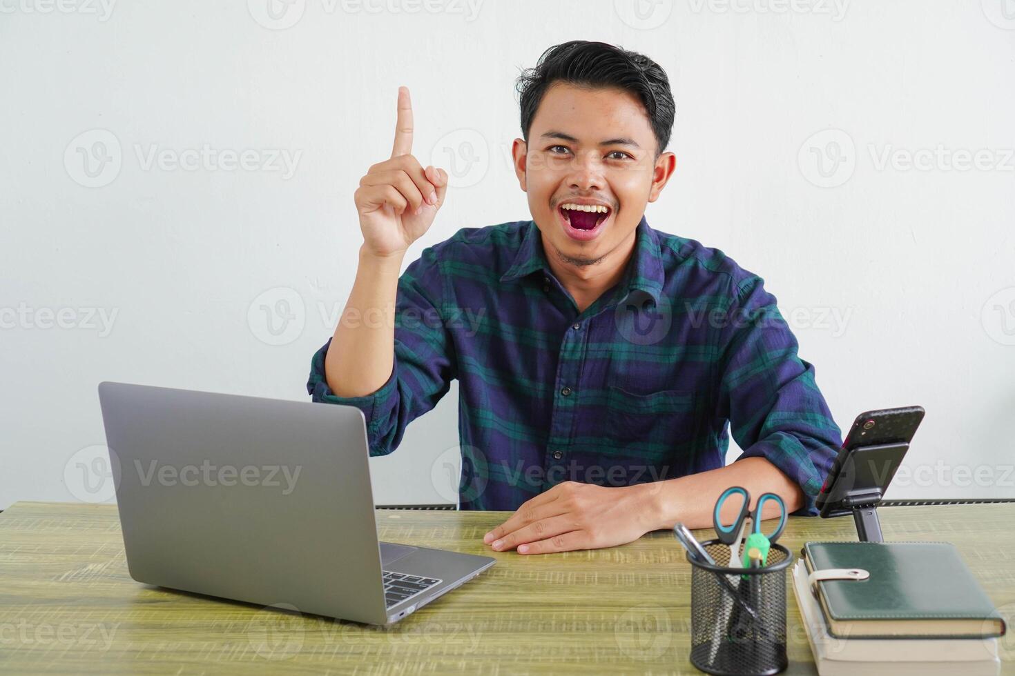 Happy young asian man sit work at wooden desk with pc laptop. Achievement business career lifestyle concept. pointing index fingers up photo