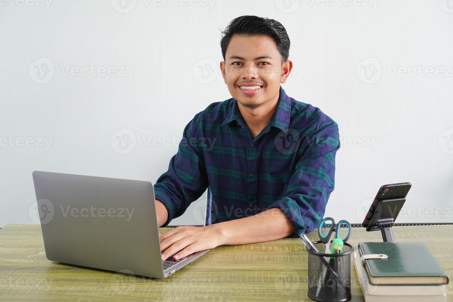 smiling young asian man in blue shirt sit work at wooden desk with pc laptop isolated on white background. Achievement business career lifestyle concept. working on project with laptop pc computer photo
