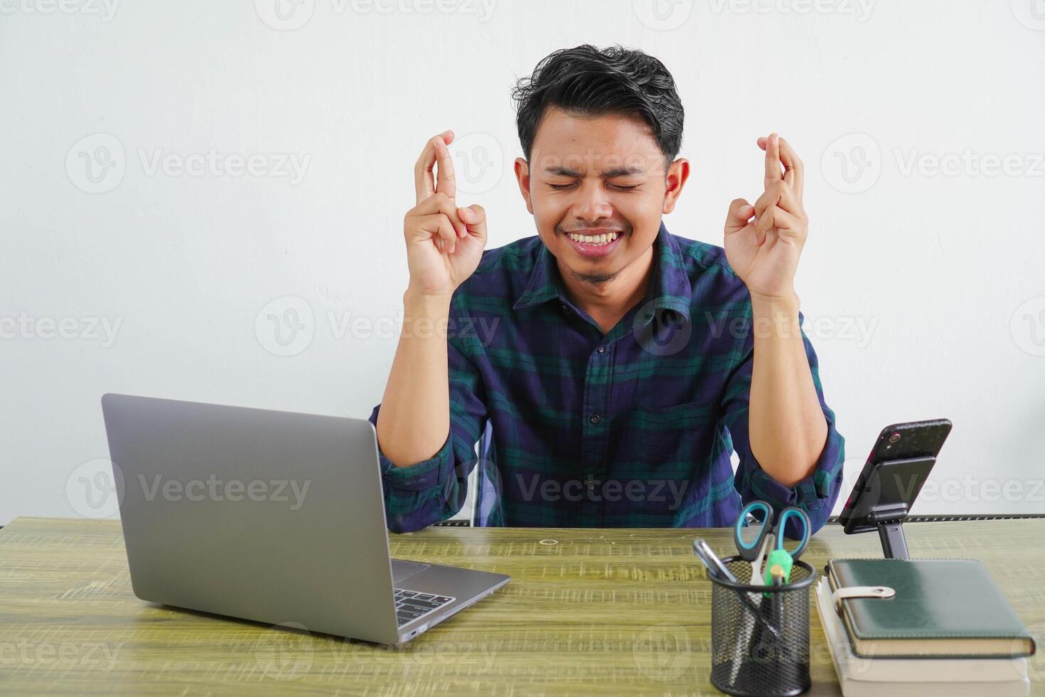 smiling young asian man sit work at wooden desk with pc laptop. Achievement business career lifestyle concept. keeping fingers crossed, eyes closed, making wish photo