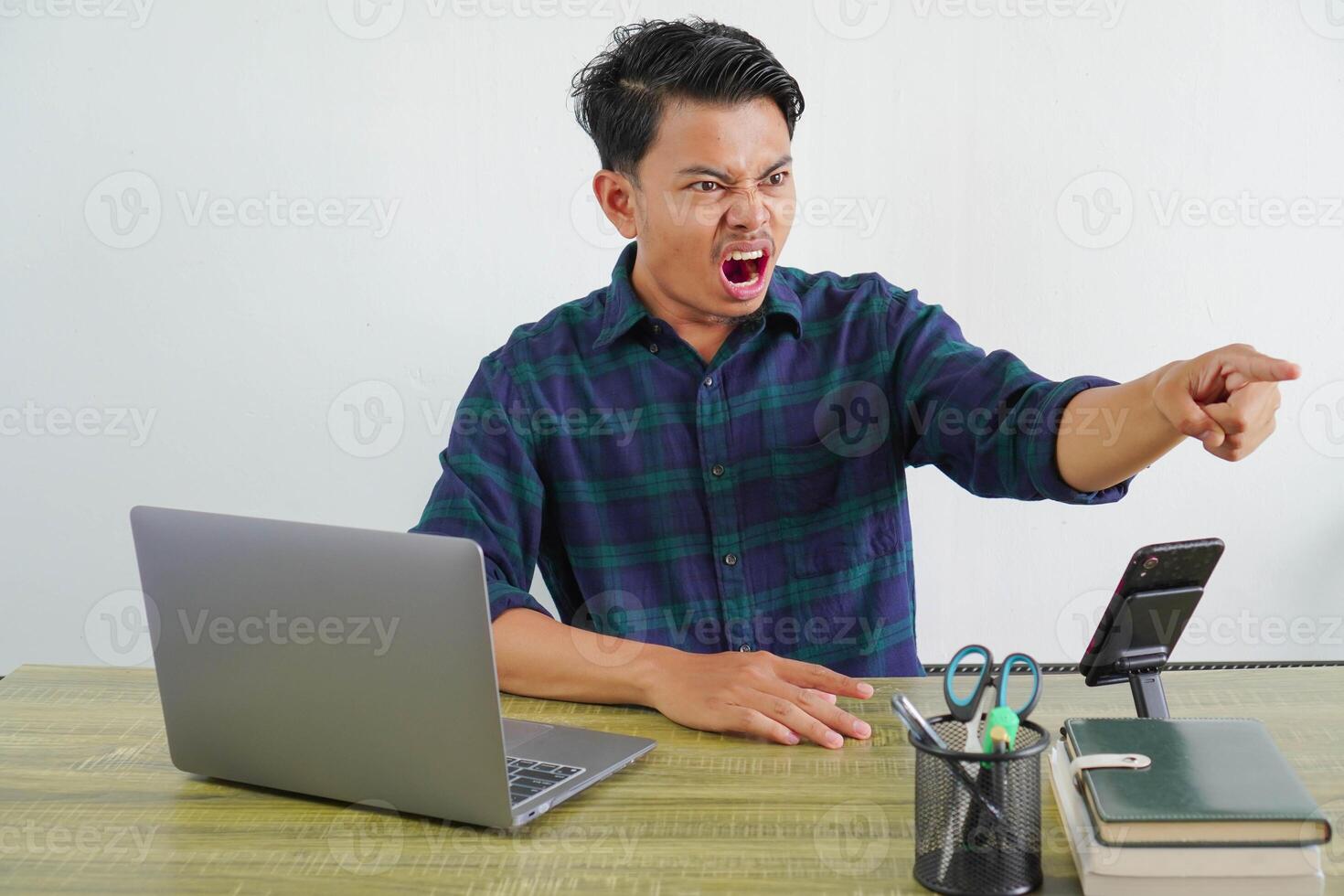Angry young asian man sit work at wooden desk with pc laptop. Achievement business career lifestyle concept. pointing index finger aside screaming swearing photo