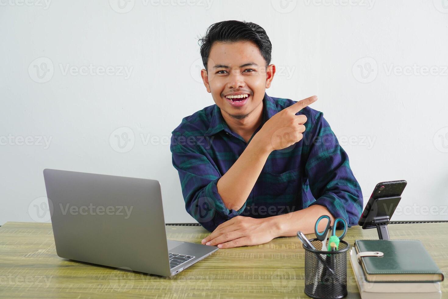 Cheerful young asian man in blue shirt sit work at wooden desk with pc laptop isolated on white background. Achievement business career lifestyle concept. pointing index finger a side up photo
