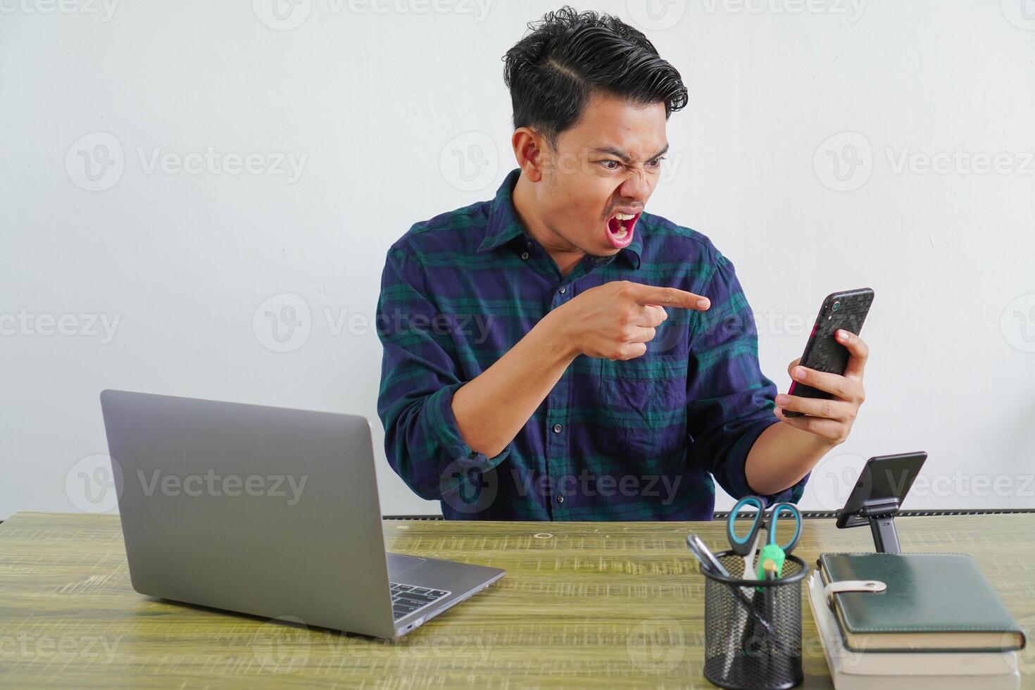 Angry young asian man sit work at wooden desk with pc laptop. Achievement business career lifestyle concept. talking and pointing on mobile phone photo