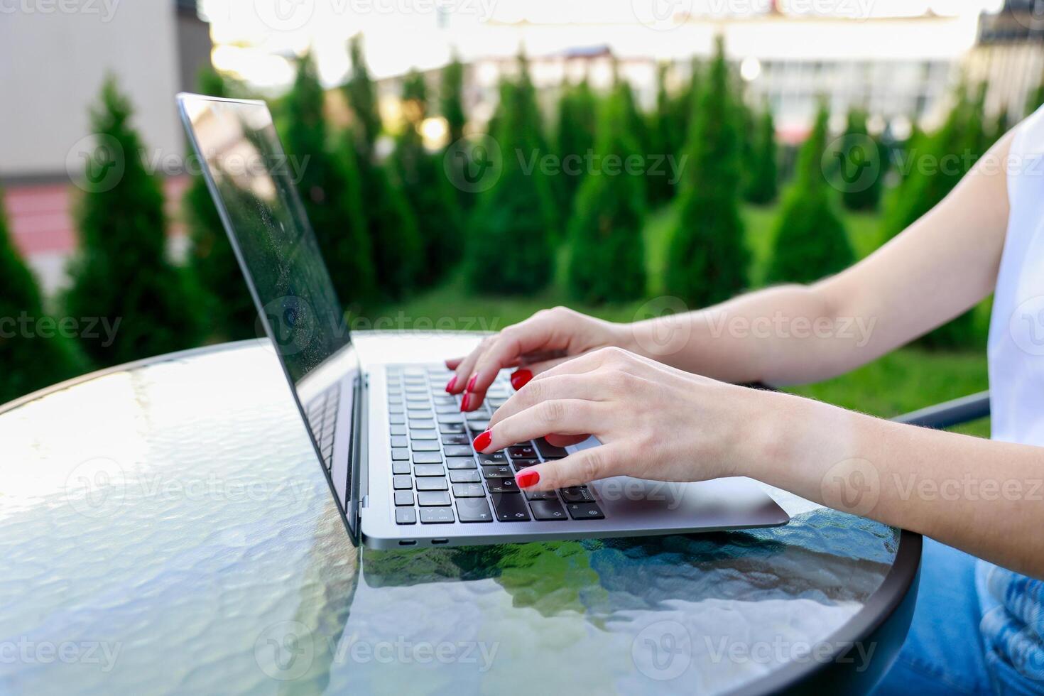 Close-up of woman hands typing on laptop outdoors. She has bright red nails, and the glass table photo