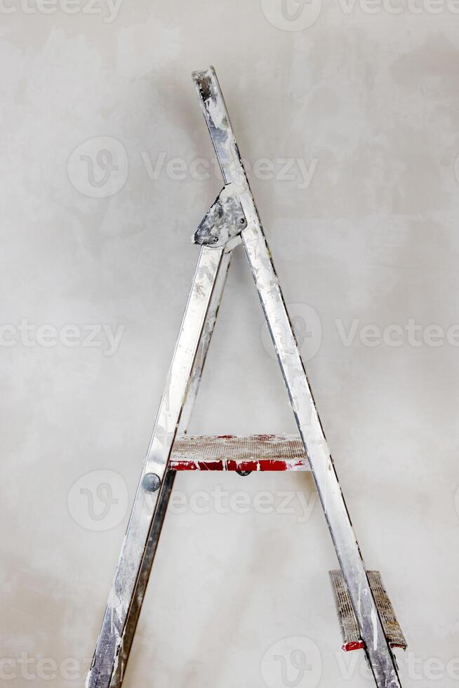 Detail of spattered paint step ladder with red step staying against the concrete unpainted wall. Home renovation DIY essential tools. Minimalistic concept. Copy space. photo
