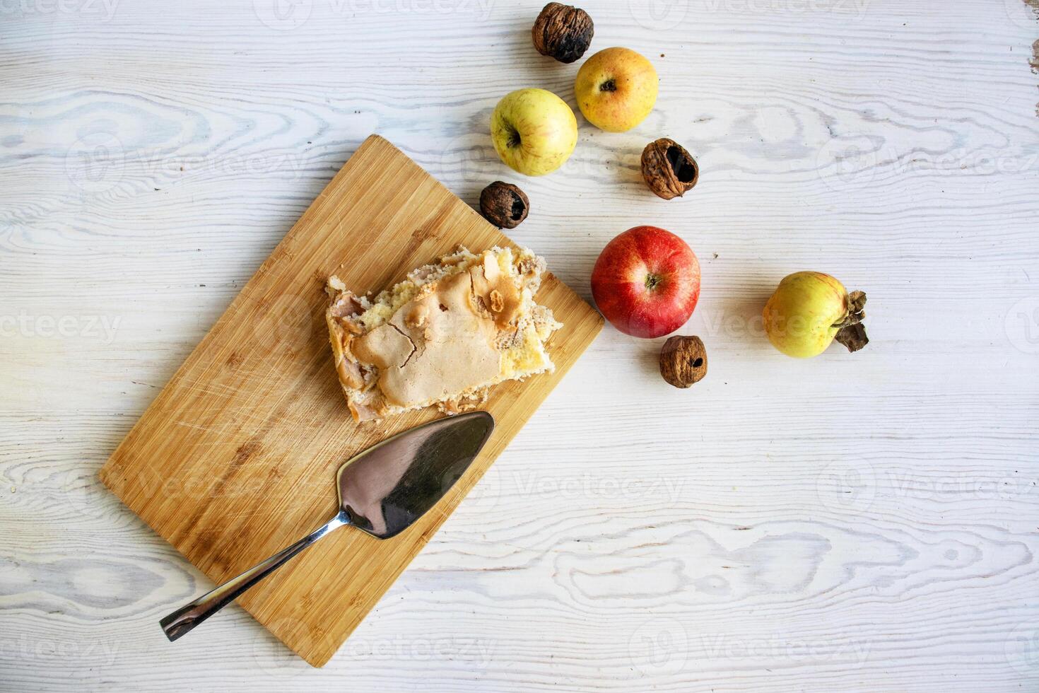 Piece of traditional thanksgiving apple cinnamon pie on the wooden cutting board on the white table, surrounded by fresh apples and walnuts. Autumn dessert. Closeup. Copy space. Top view photo