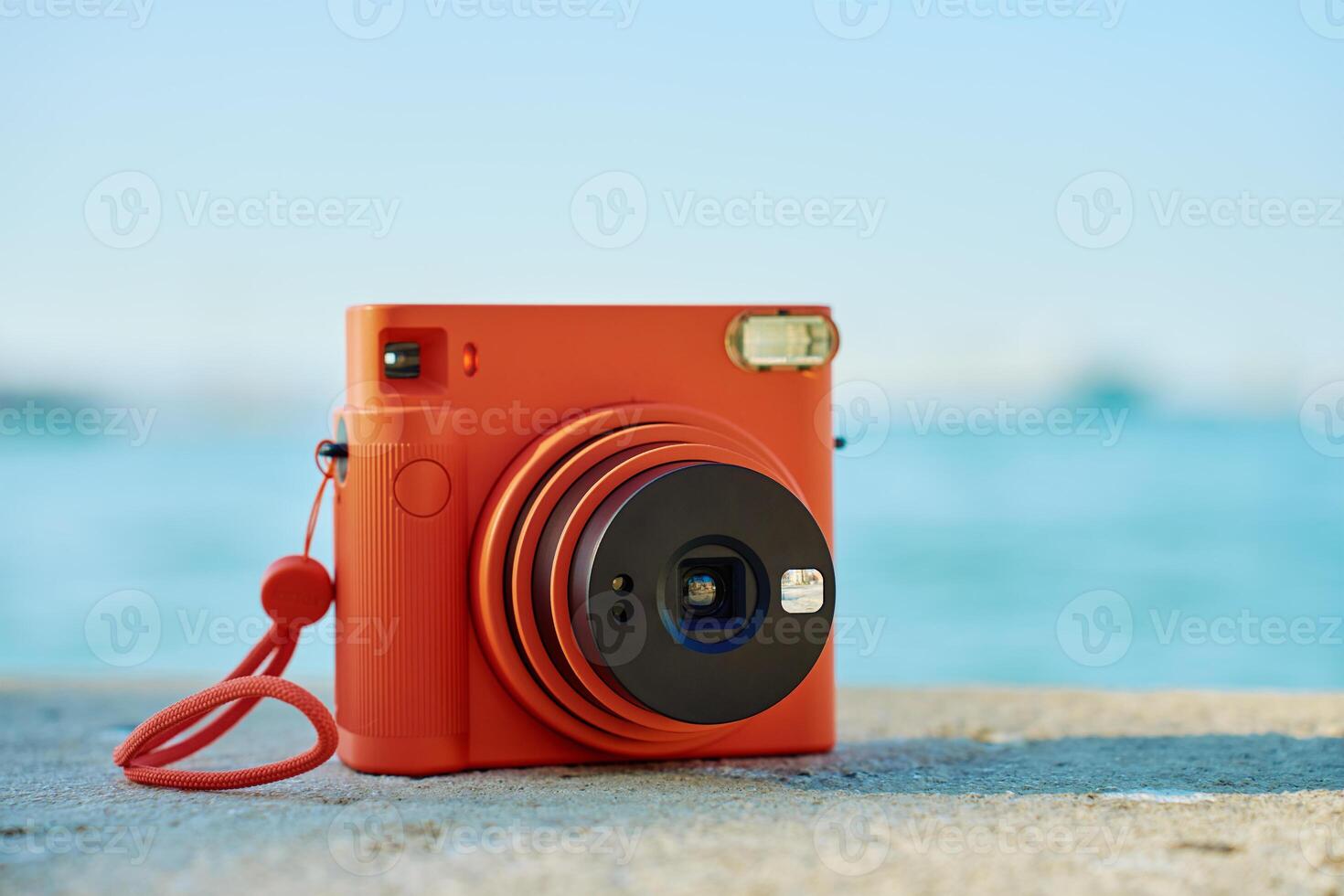Vintage orange instant camera on rock by seaside with blurred coastal town in background. Summer travel concept. Equipment for capturing moments photo